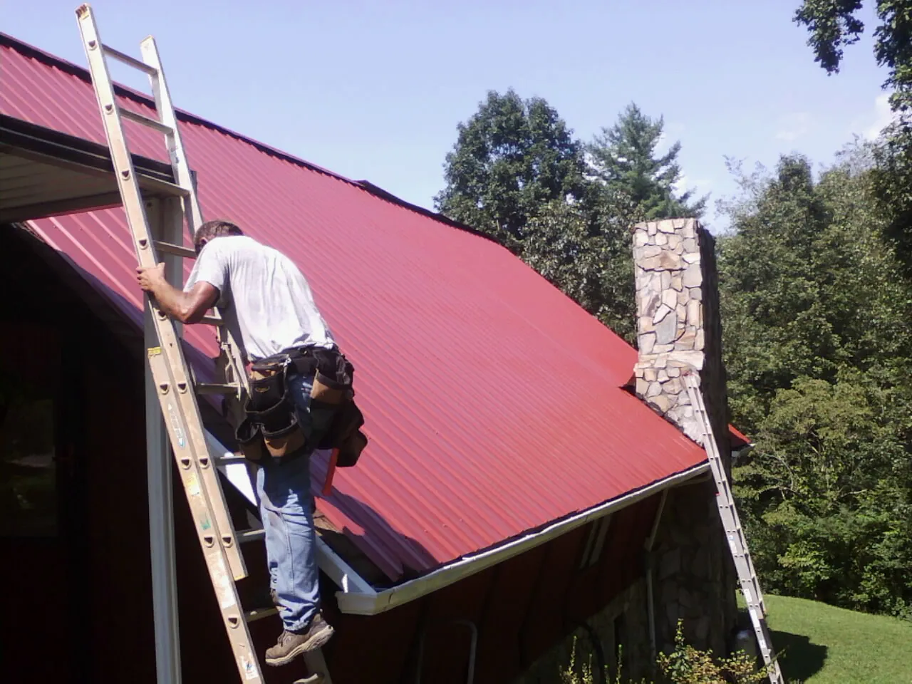 Roofer on a ladder beside a red metal roof and stone chimney — Soffit & Fascia Repair by Roofing Brothers in Mulino